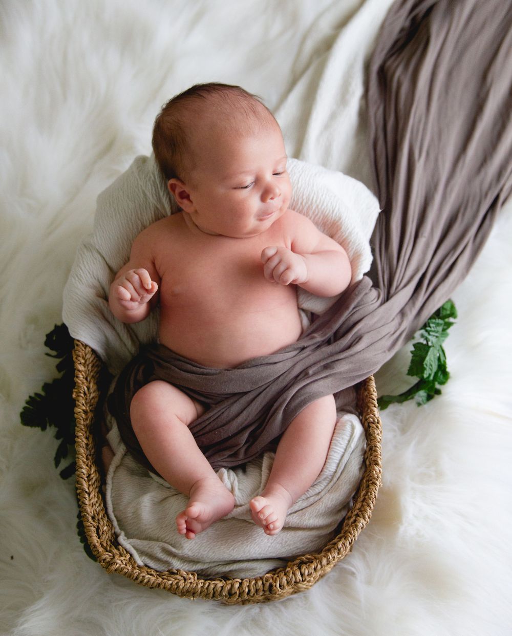 Newborn baby rests peacefully in woven basket with gray fabric draping.