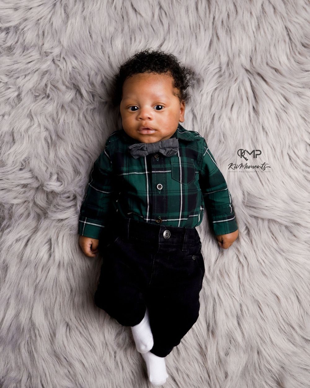 Baby in plaid shirt and dark pants poses on gray fur background.