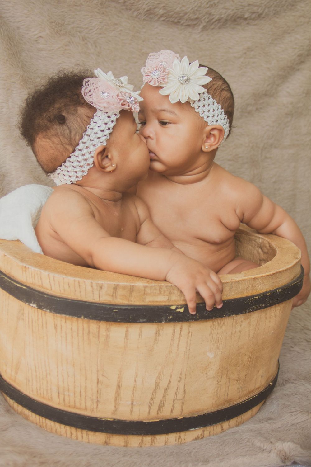 Two babies wearing lace headbands sit in wooden barrel during portrait session.