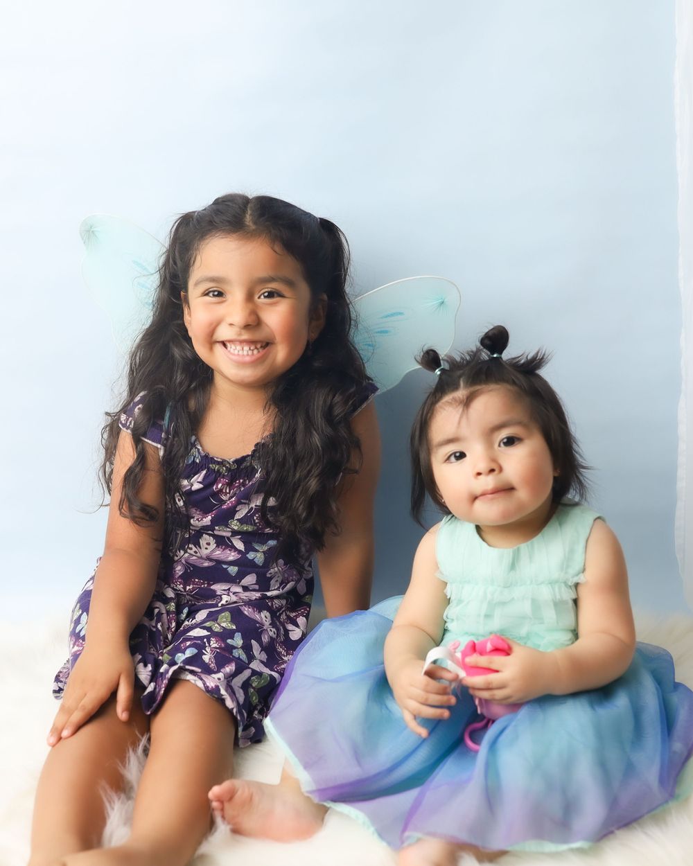 Two small children wearing fancy dresses smile while sitting together against a light background.