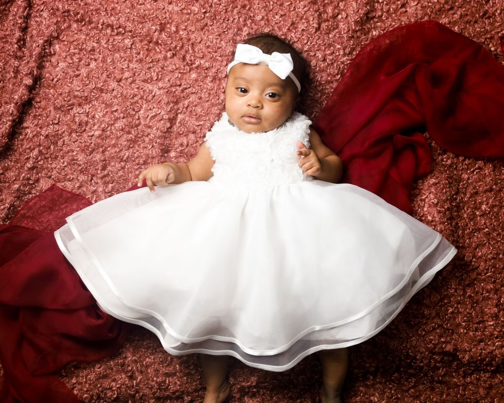 Baby in white christening dress poses on red textured backdrop.