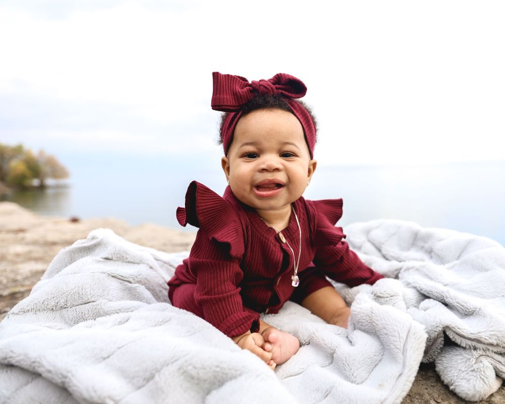 Baby in burgundy ruffled outfit smiling on blanket outdoors.