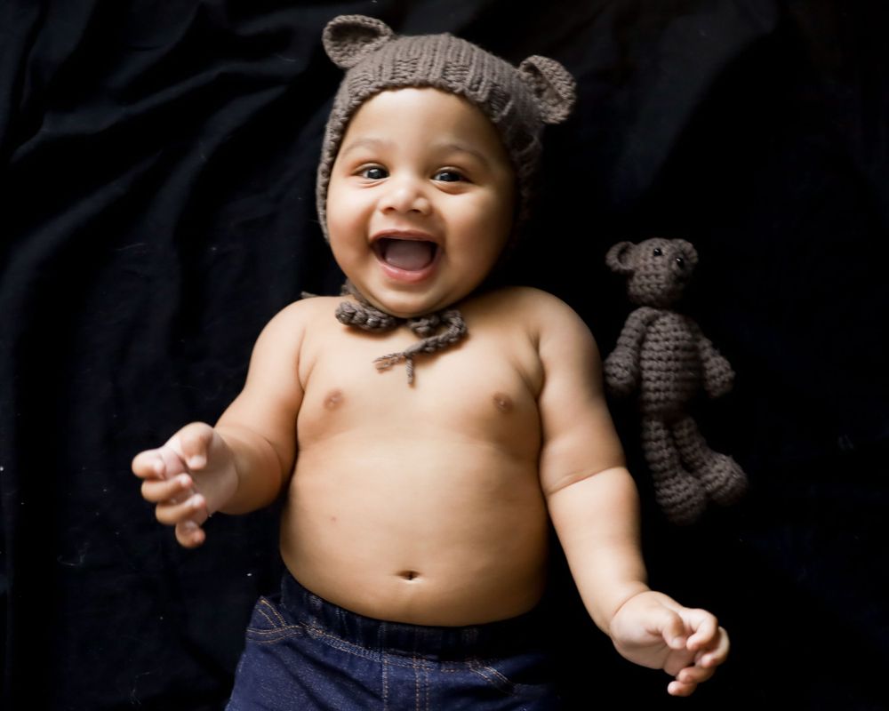 Baby wearing knit bear hat smiles joyfully with stuffed animal on black background.