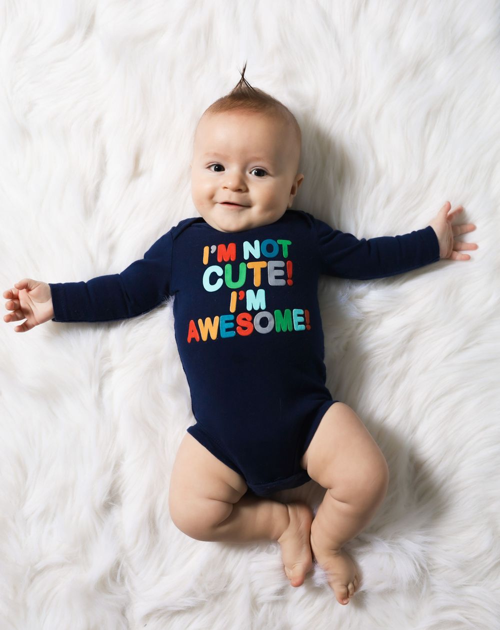 Baby wearing navy onesie with colorful text lying on white fur rug.