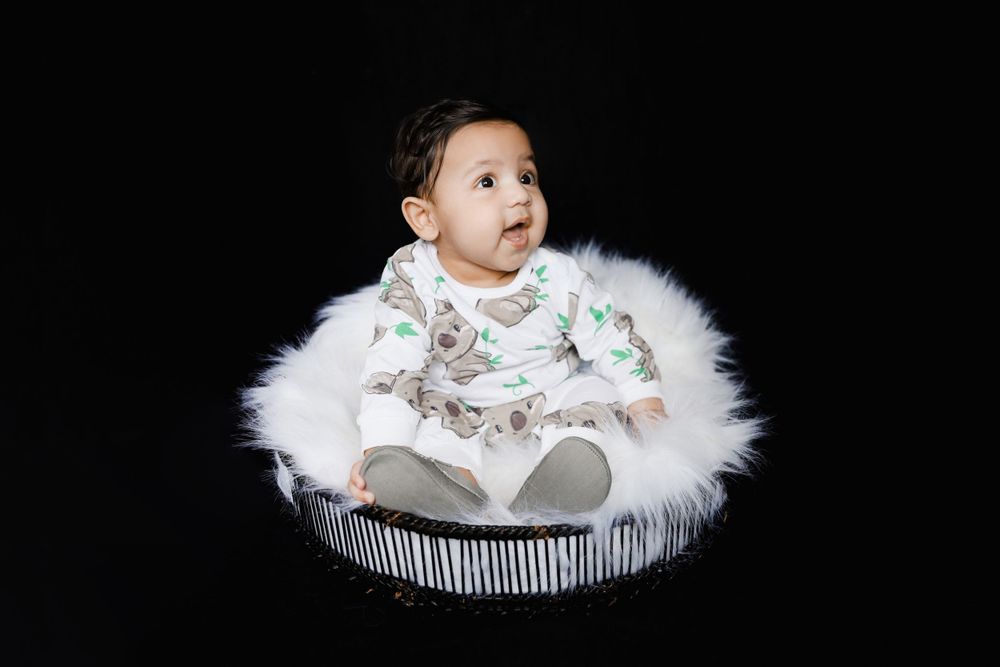 Baby in floral outfit sits on white fur cushion against dark background.