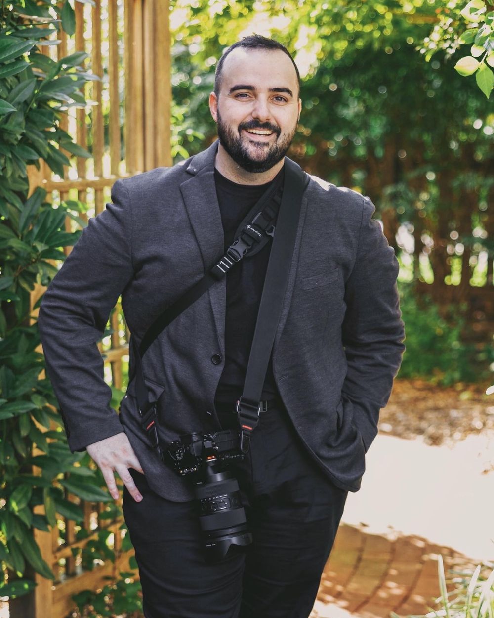 Professional photographer in dark attire with camera gear standing in a garden setting.