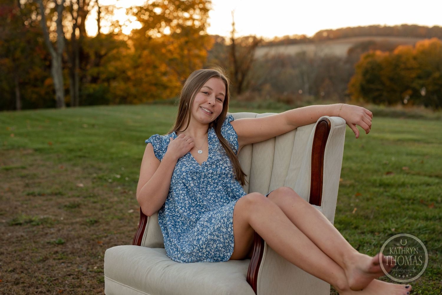 Person lounges in beige armchair outdoors wearing blue dress during golden hour.