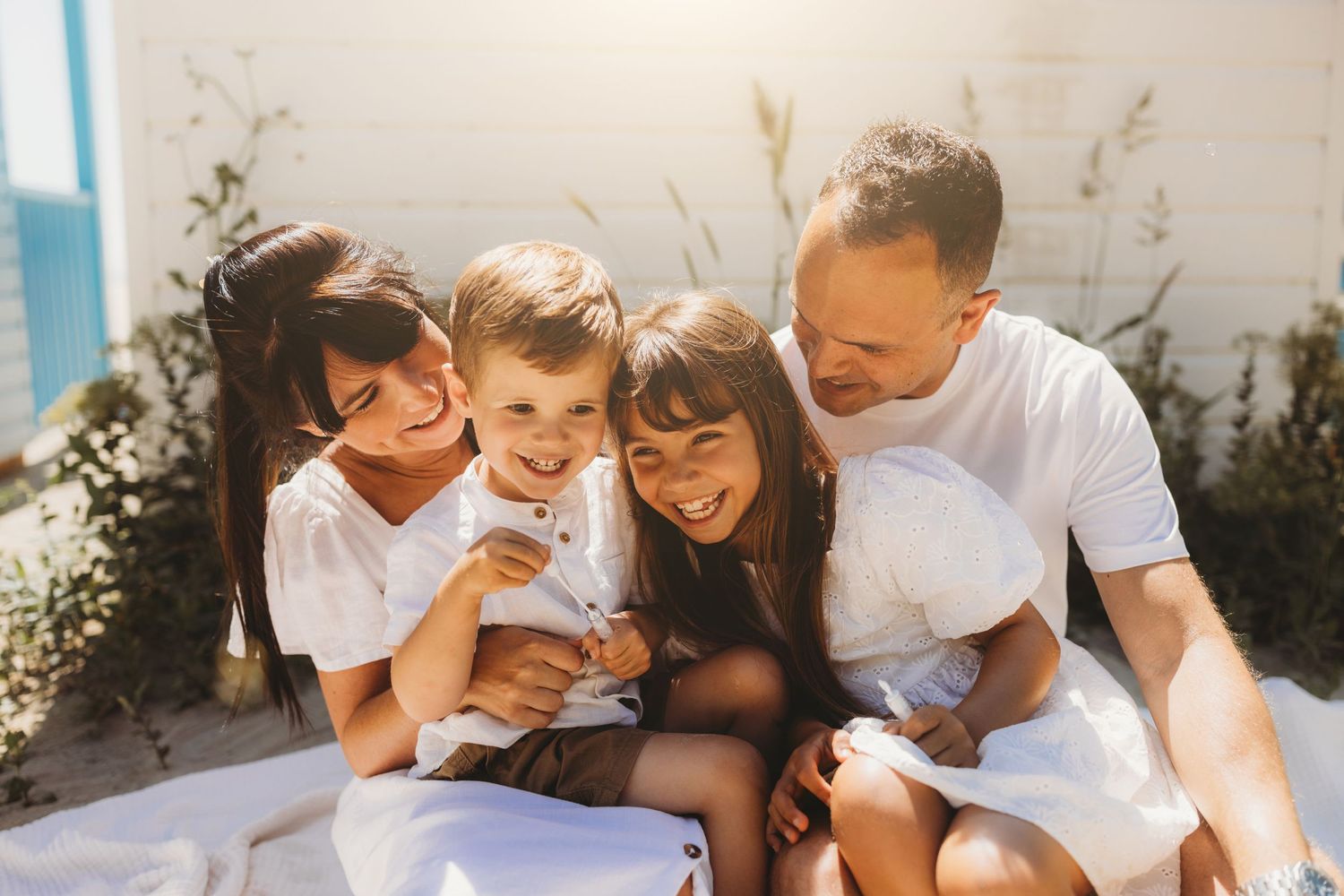 Family wearing white clothes sits together on blanket outdoors during golden hour, sharing joyful laughter together.