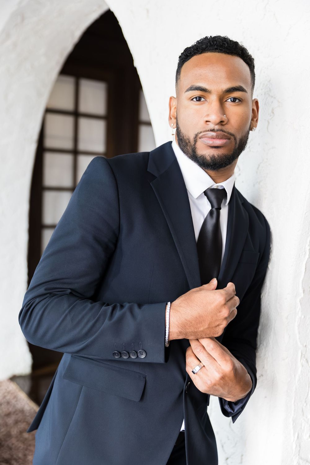 Confident Black groom in navy suit and black tie leaning against white archway wall.