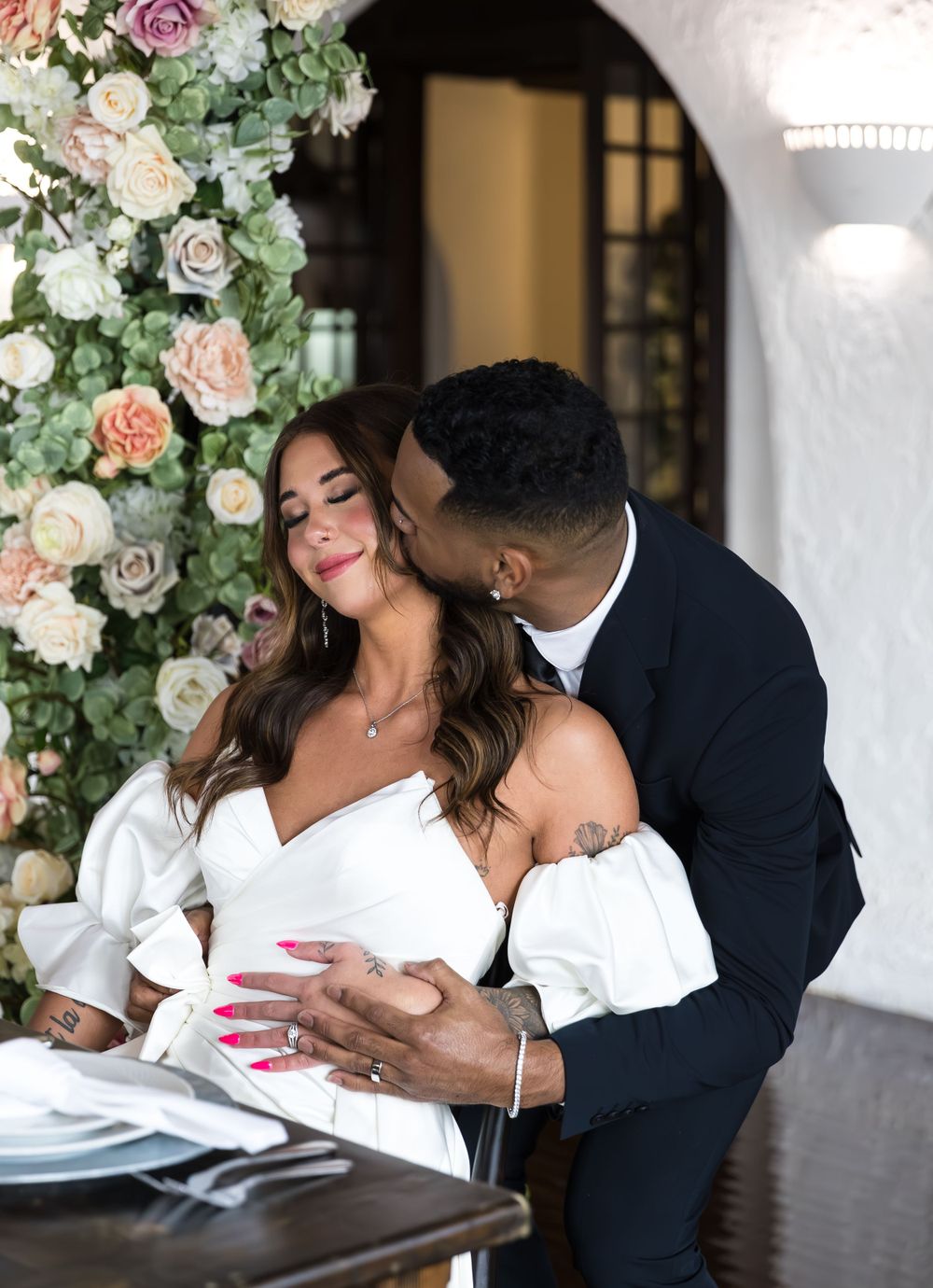 Groom in dark suit kissing bride in white off-shoulder dress beside a floral arrangement at wedding.