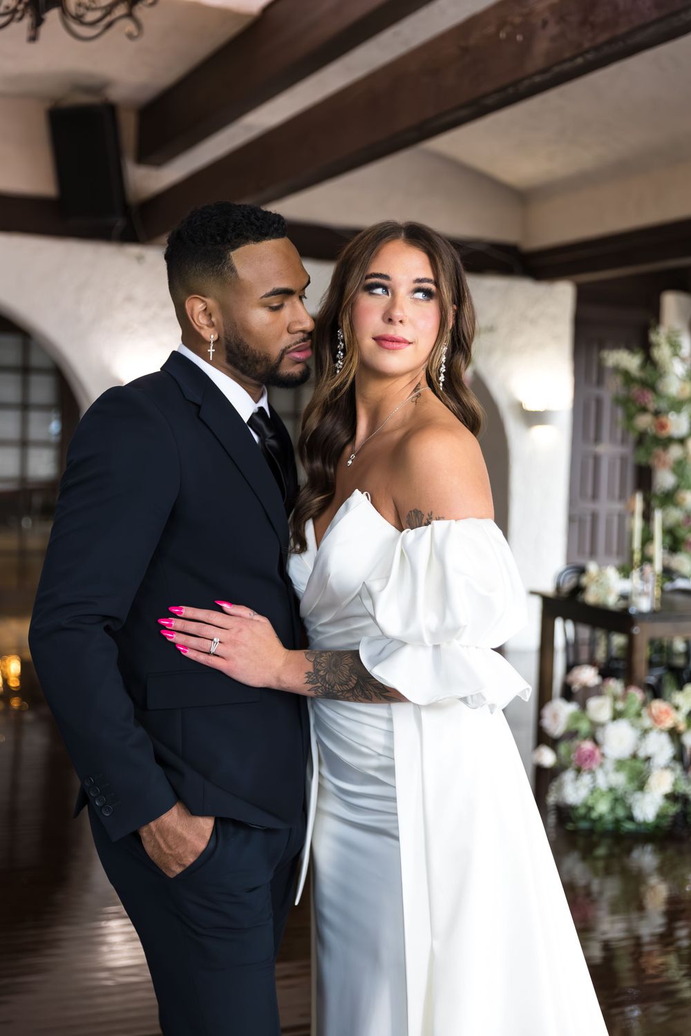 Elegant interracial wedding couple posing indoors, bride in off-shoulder white gown with groom in dark suit.
