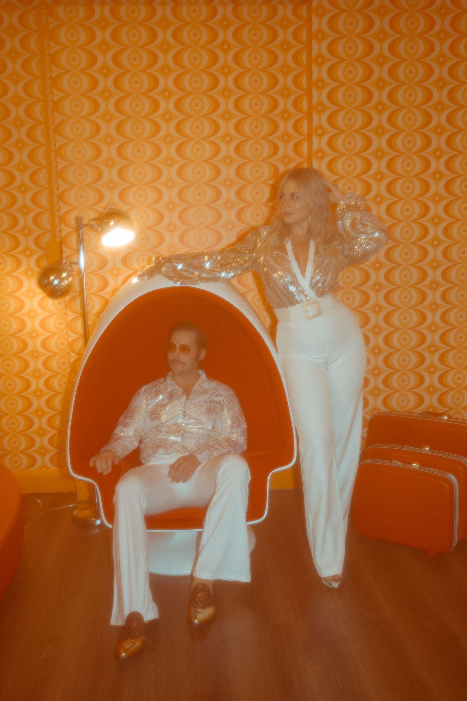 Retro interior scene with orange patterned wallpaper, red chair, and two people in white outfits illuminated by warm lighting.