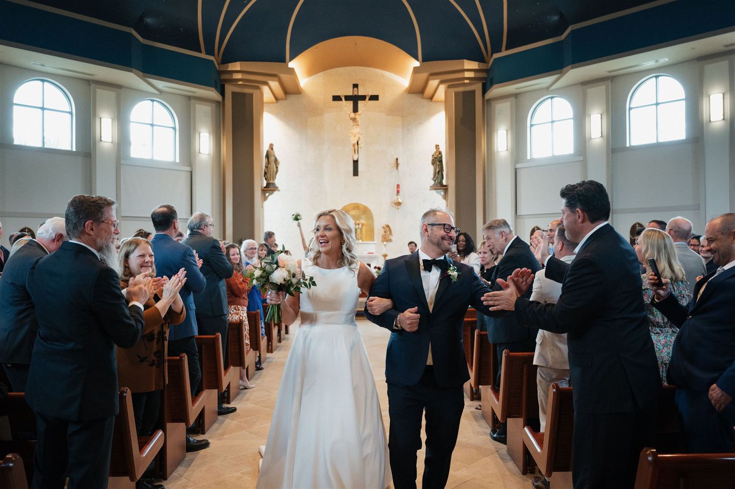Wedding guests applaud as newlyweds exit down the aisle in a church with arched ceilings.