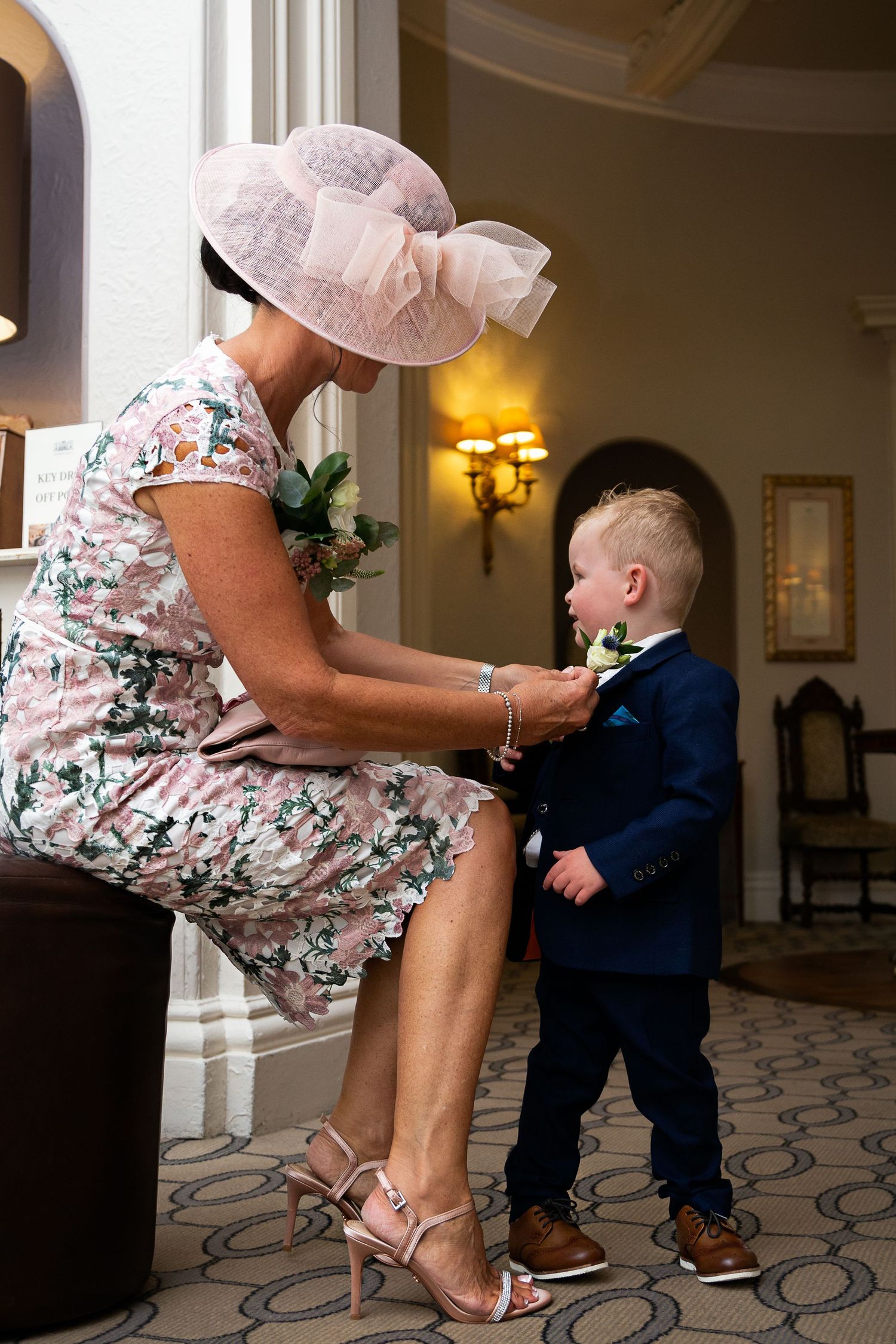 A sweet interaction between an elegantly dressed woman in a pink hat and small boy in a navy suit.