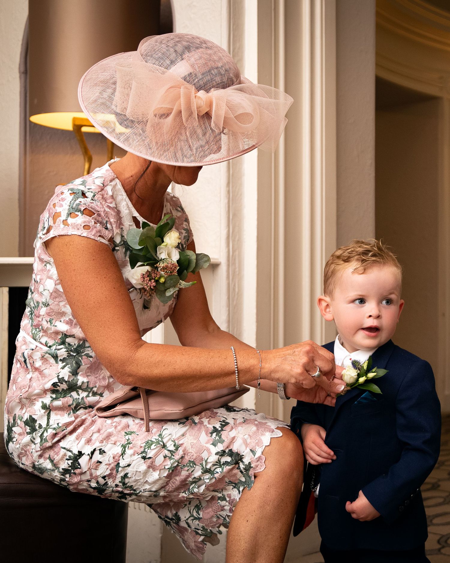 A touching moment at a formal event as a woman in a floral dress and pink hat adjusts a young boy's boutonniere.