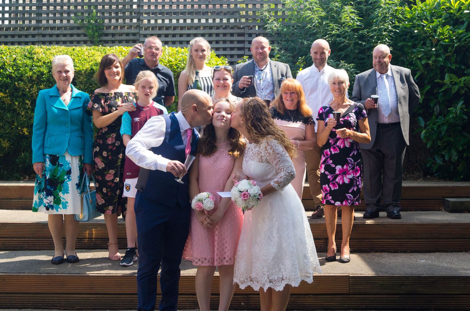 A multi-generational wedding party poses on outdoor wooden steps surrounded by greenery.