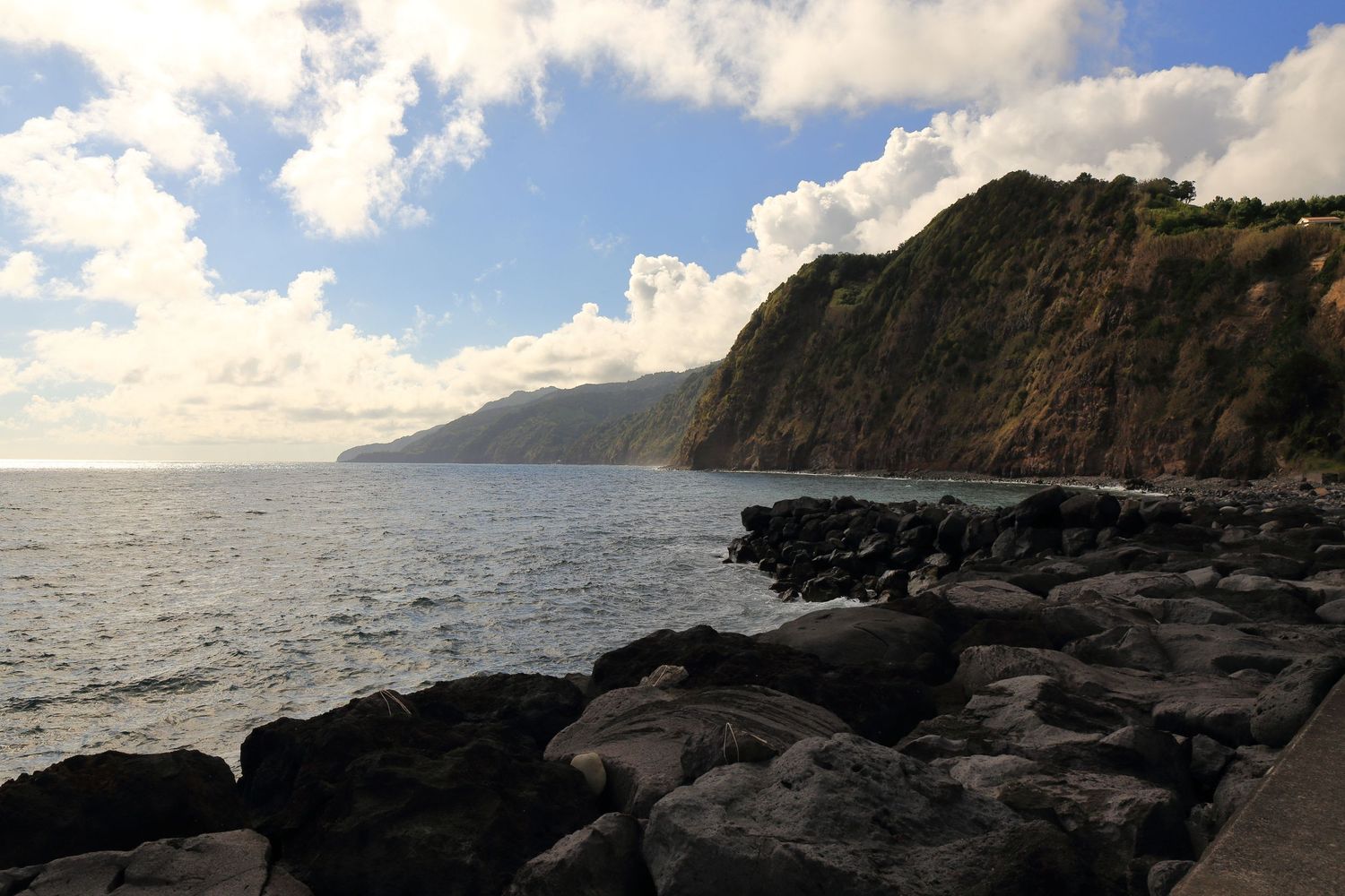 Rocky coastline with steep cliffs meets the ocean under bright sunlight and blue skies.