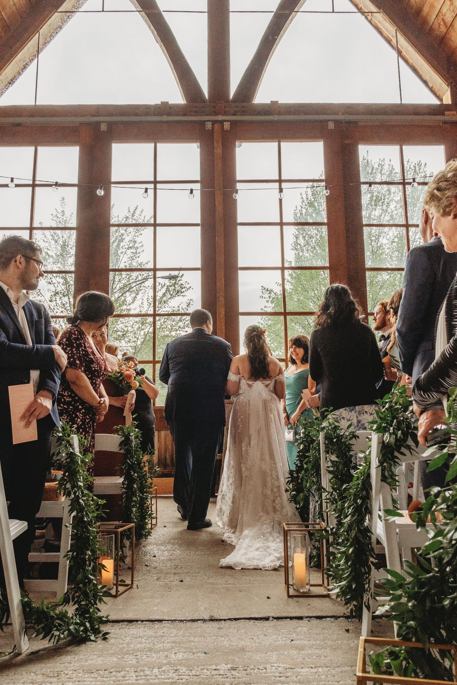 Wedding ceremony in rustic wood beam venue with lantern-lined aisle and greenery decorations.