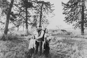 Black and white photo of a family of four embracing among tall pine trees in a field.