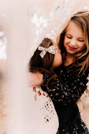 Two young girls hugging and smiling, one wearing a floral bow in her hair, soft bokeh background.