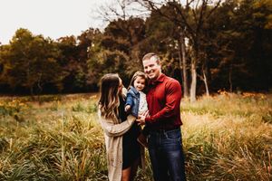 Family of three posing in autumn field with colorful fall foliage backdrop.