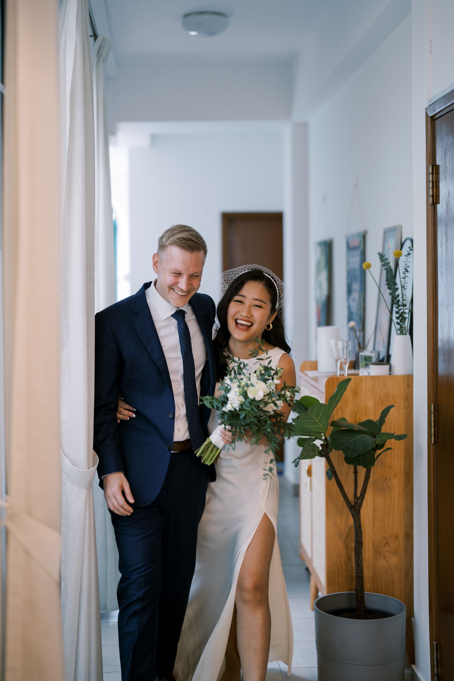 Newlyweds walking and laughing together in a bright white hallway.