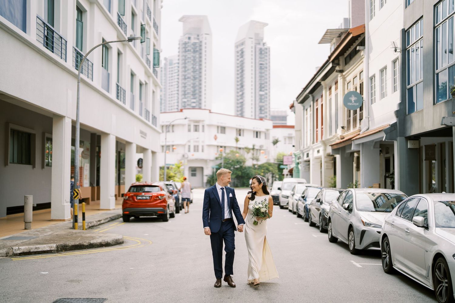Wedding couple walking together on a city street lined with parked cars and high-rise buildings.