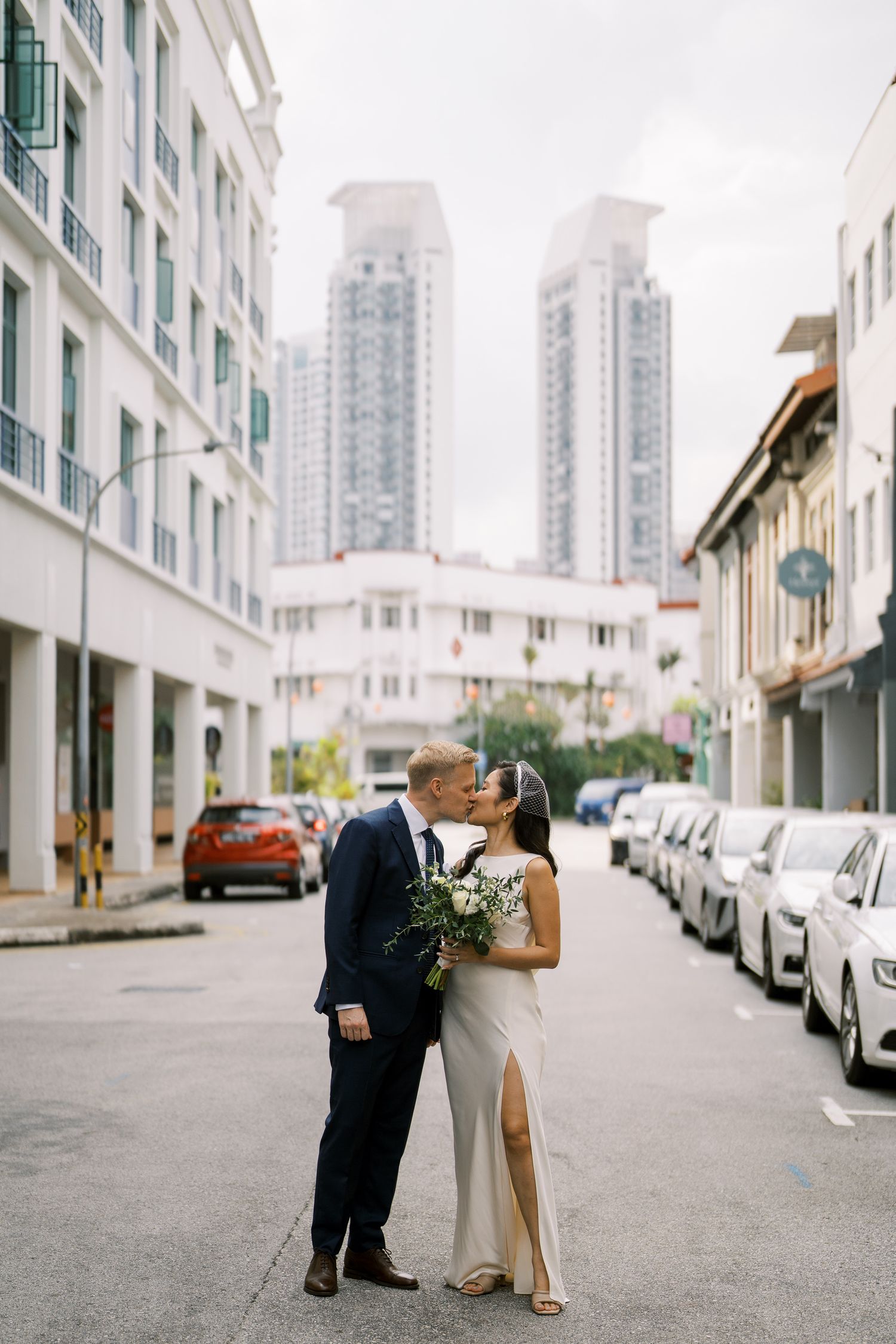 A couple in formal attire share intimate moments on a city street lined with white buildings and parked cars.