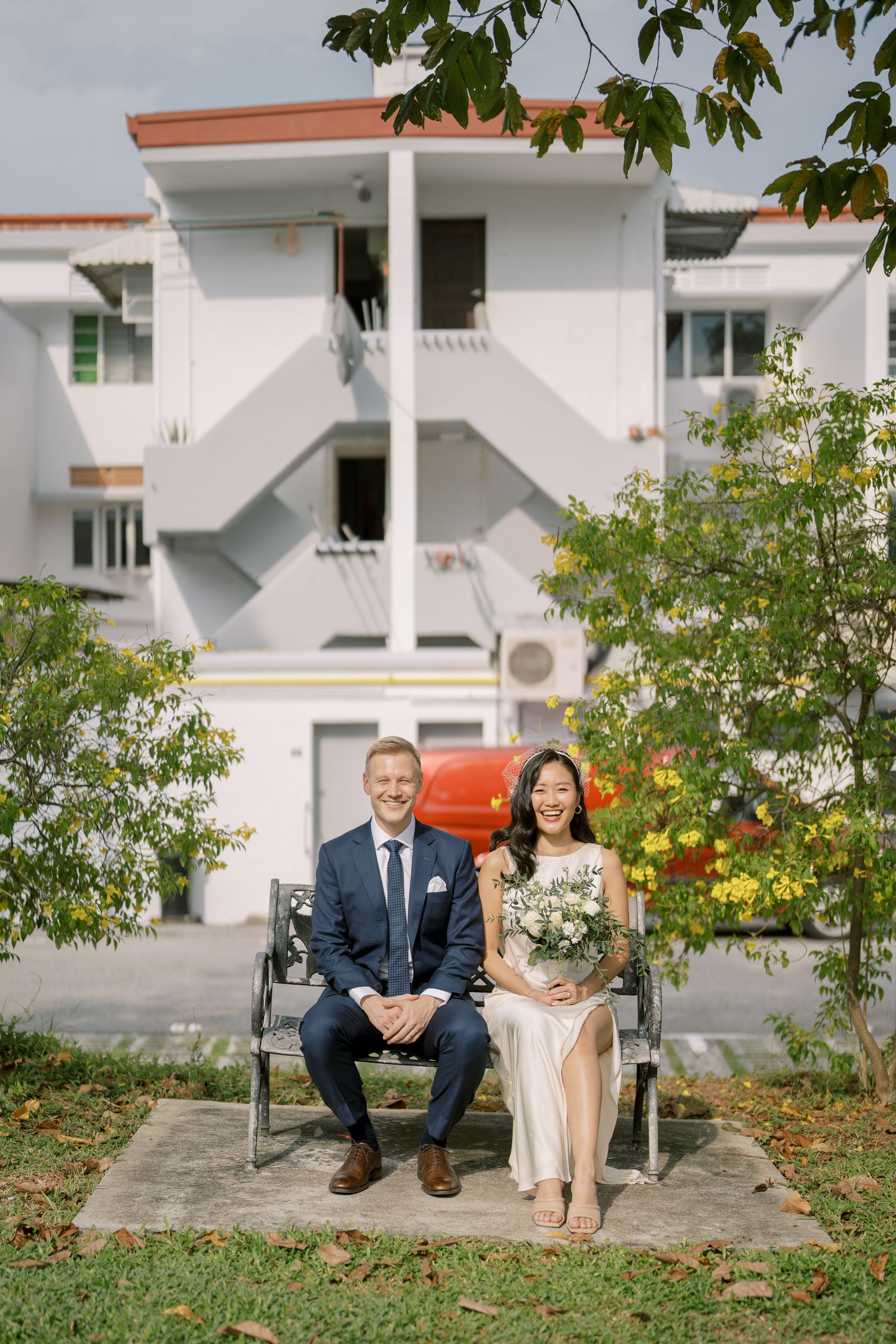 A couple sits together on concrete steps surrounded by greenery and white buildings.