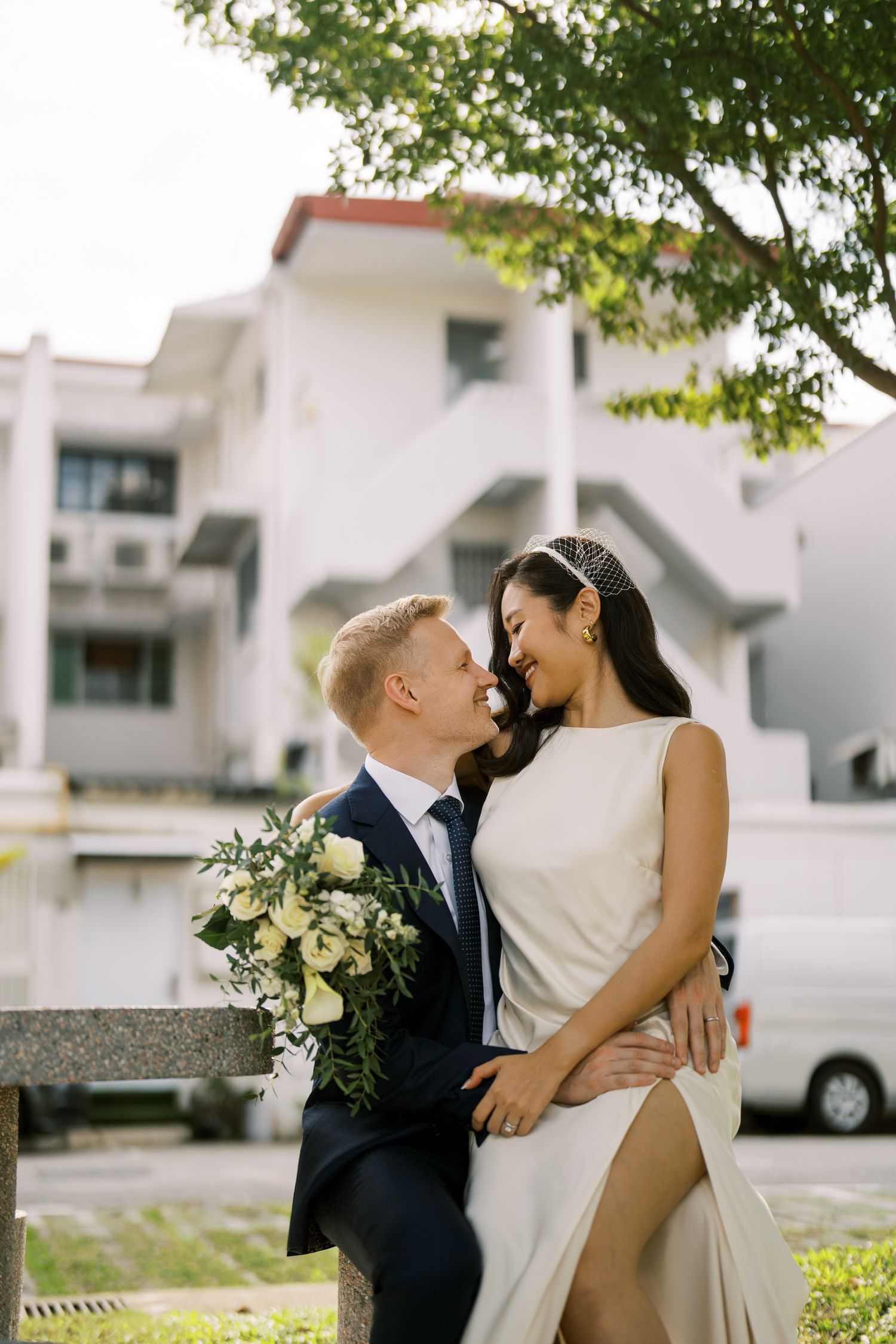 A tender embrace between a couple in formal wear near white buildings and trees.