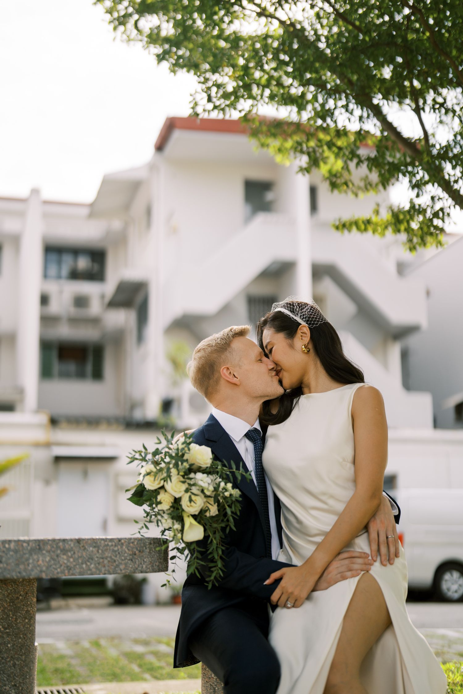 A couple shares romantic moments in an outdoor setting with white buildings and trees.