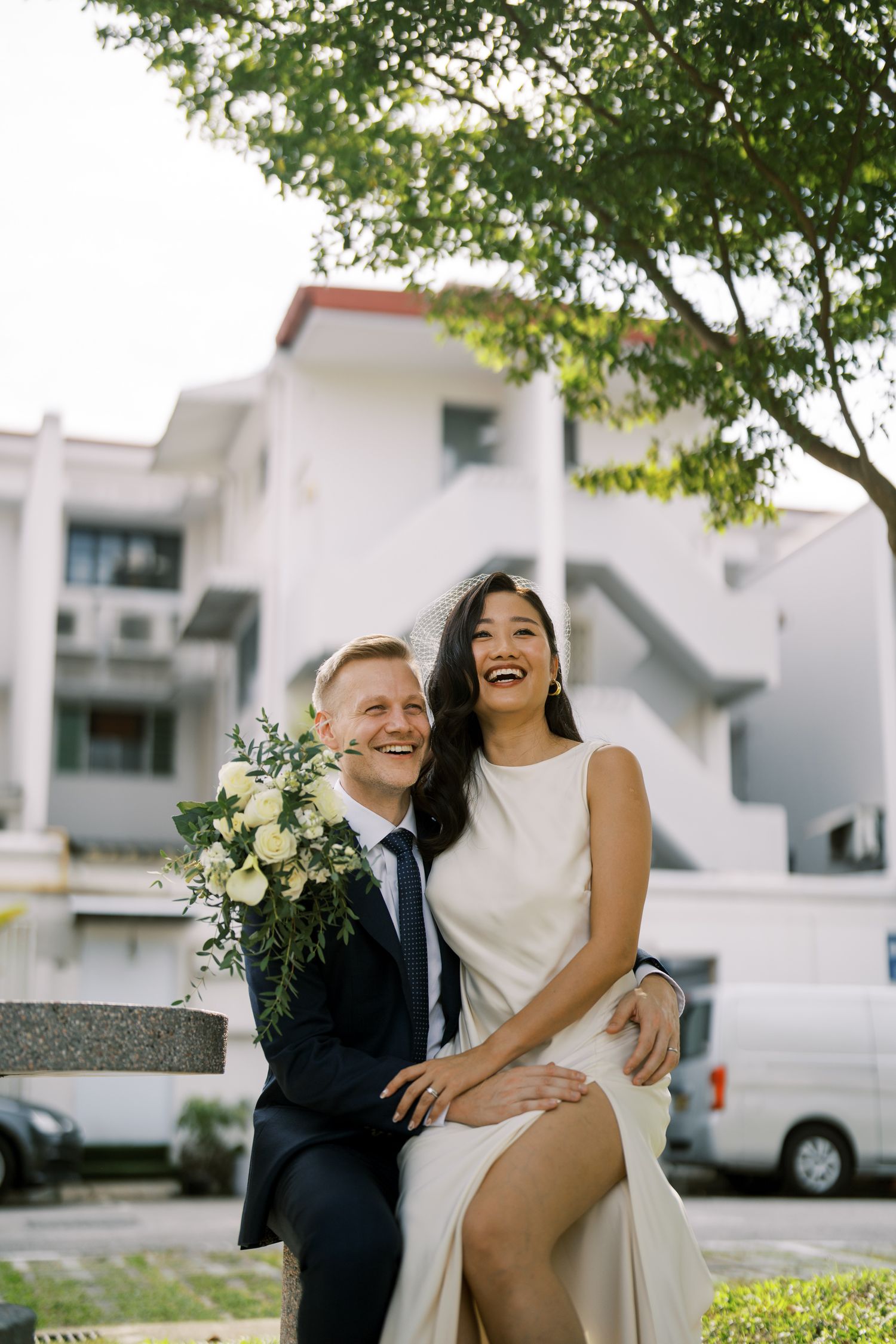 A joyful couple laughs together during their wedding photoshoot near white buildings.
