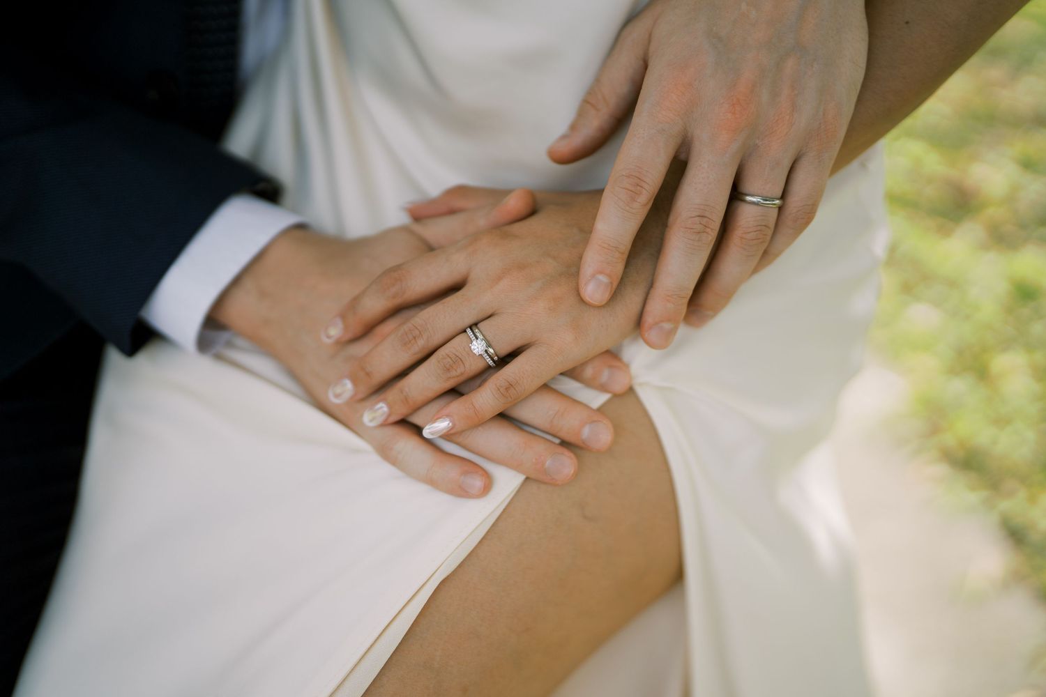 Close-up detail of hands with wedding rings against a white dress background.