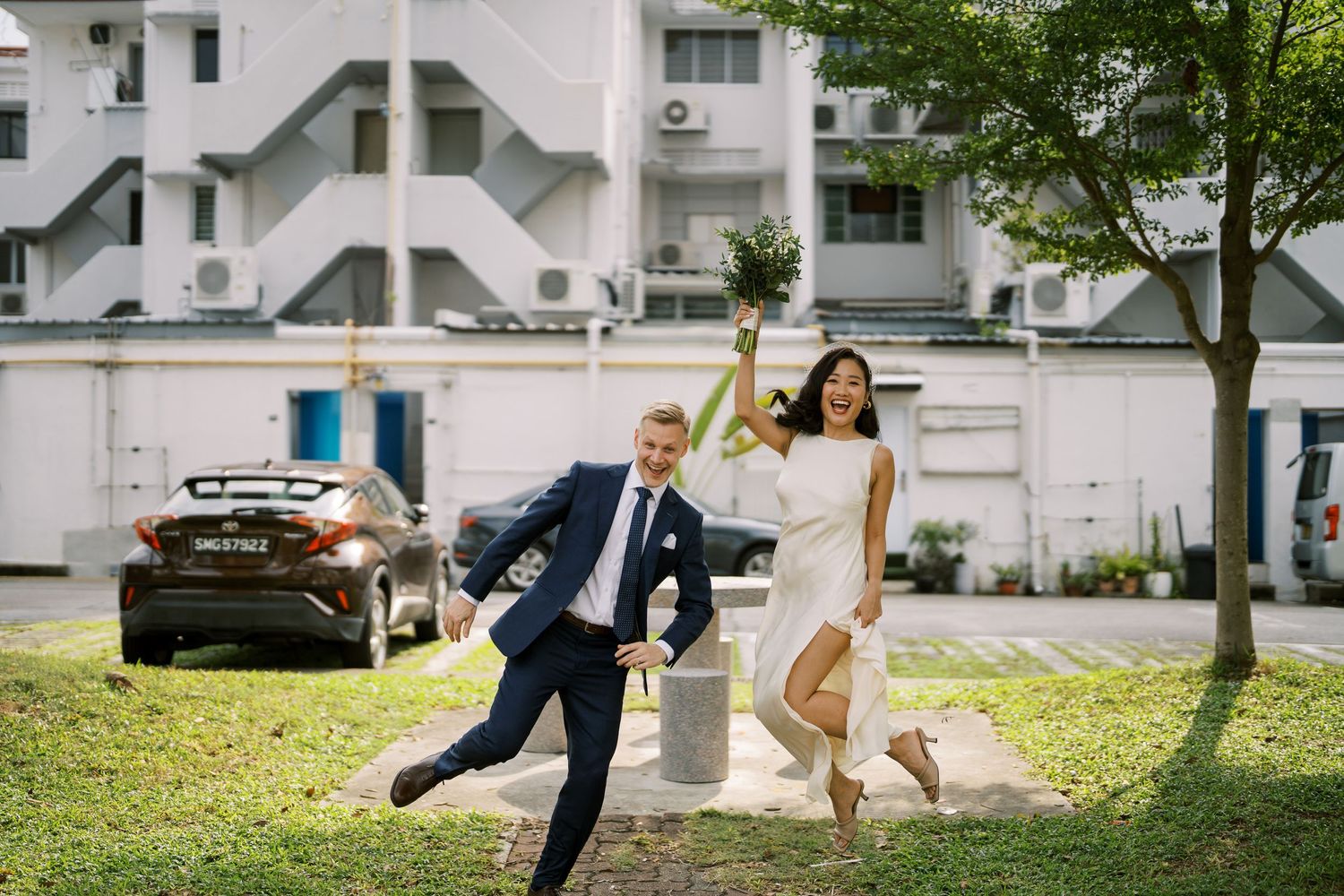 Playful wedding couple running and dancing together on grass near white buildings.