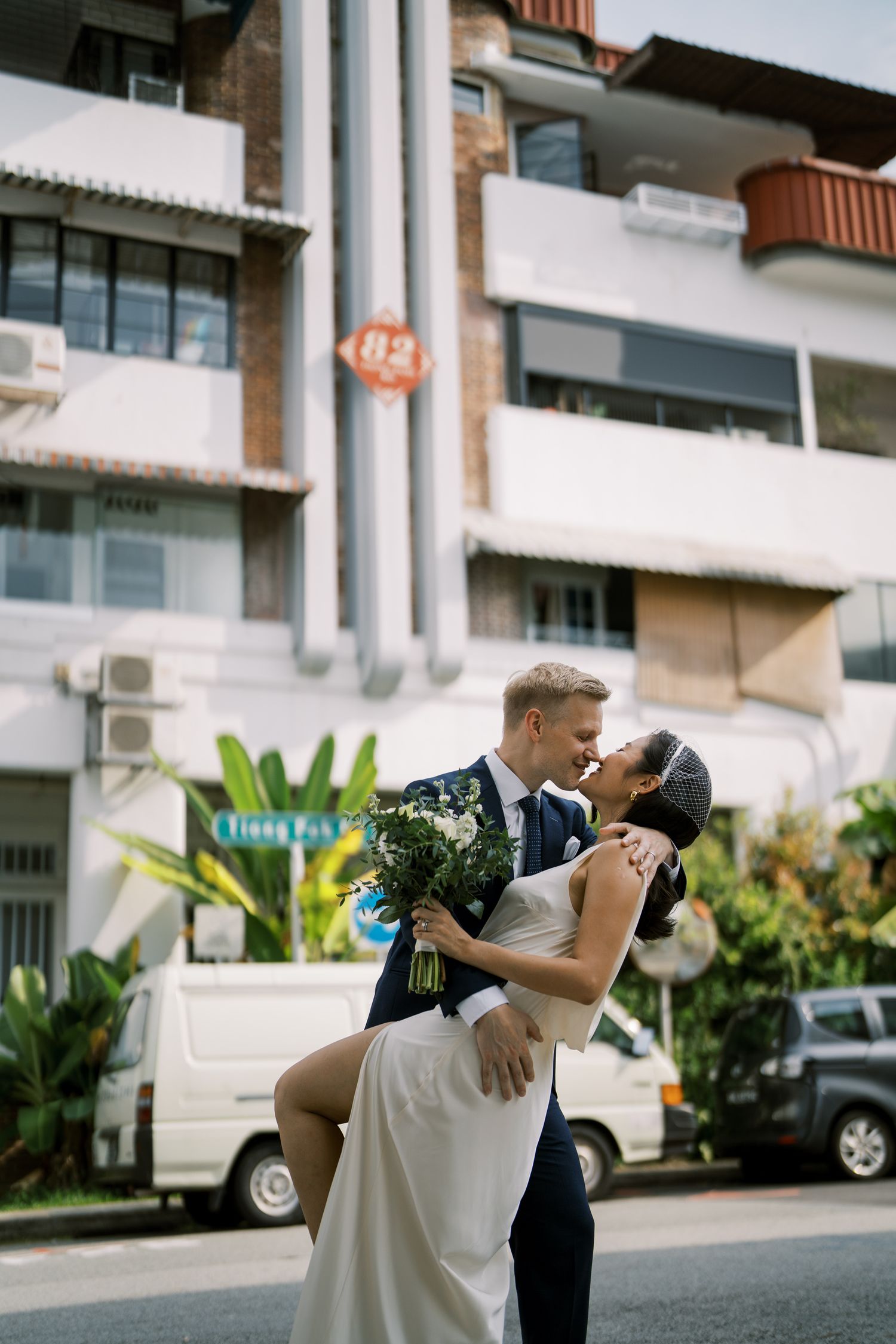 A romantic dip pose between a couple in formal attire outside a modern building with tropical landscaping.