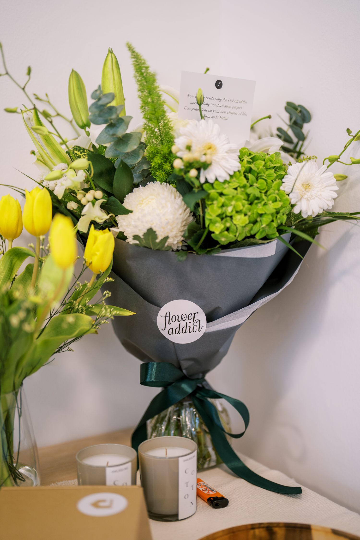 Fresh bouquet featuring yellow tulips and white flowers wrapped in dark paper with ribbon.