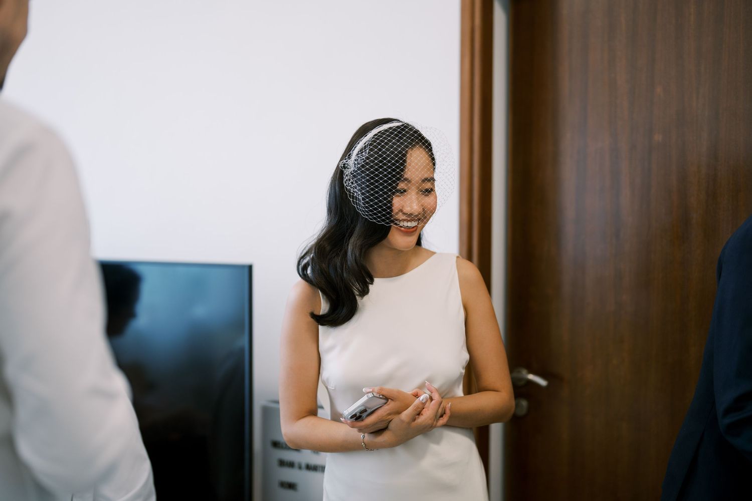 A bride wearing a white dress and headpiece smiles joyfully during her wedding preparations.