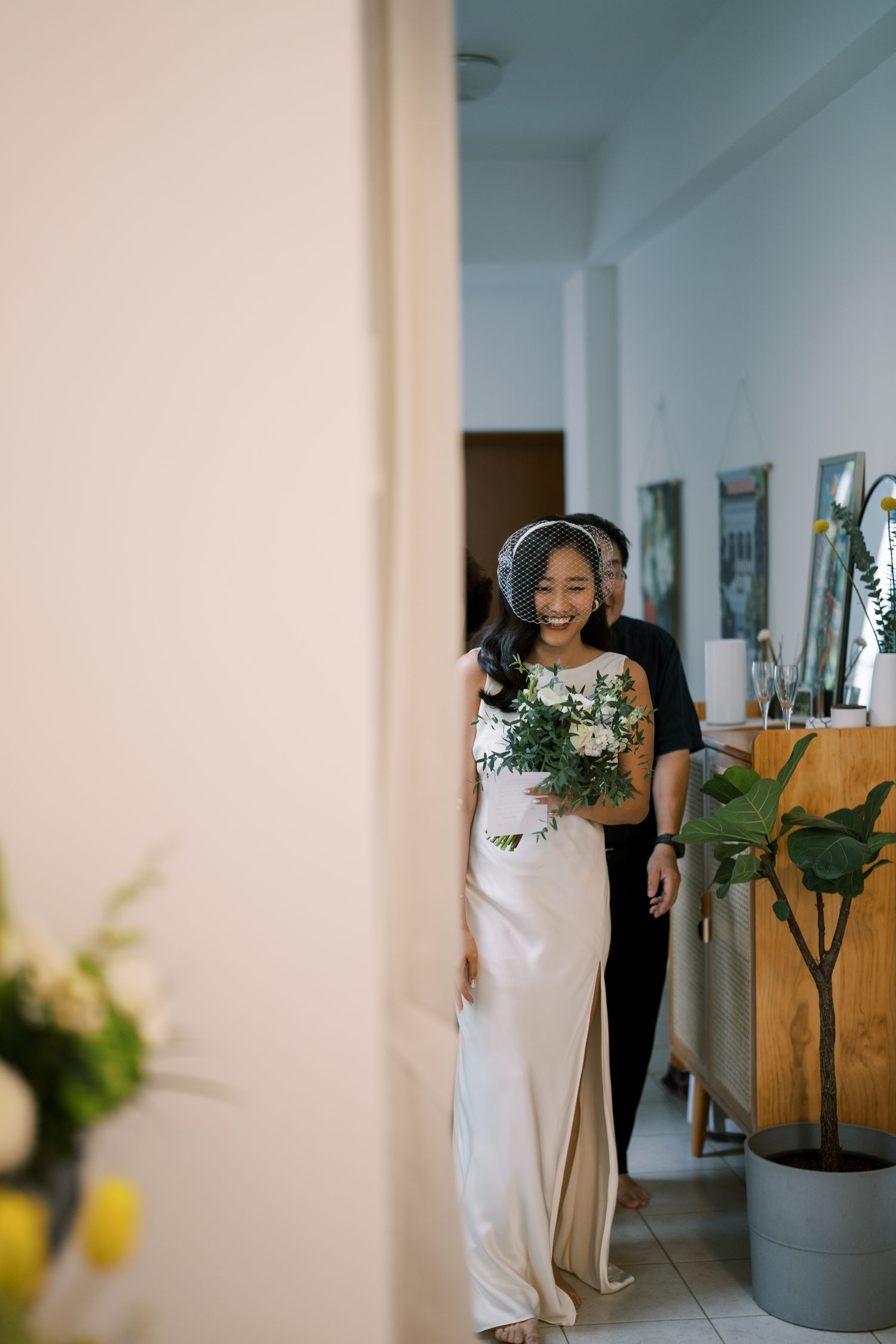 A bride in an elegant white gown walks down a hallway carrying a green and white bouquet.