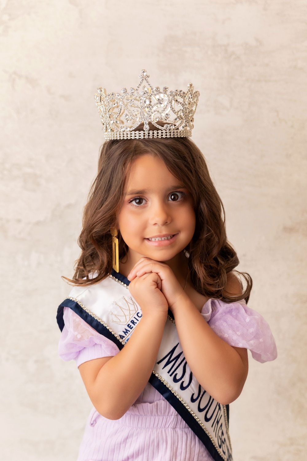 A beauty pageant contestant wearing a crown and lavender dress poses with clasped hands against a light background.