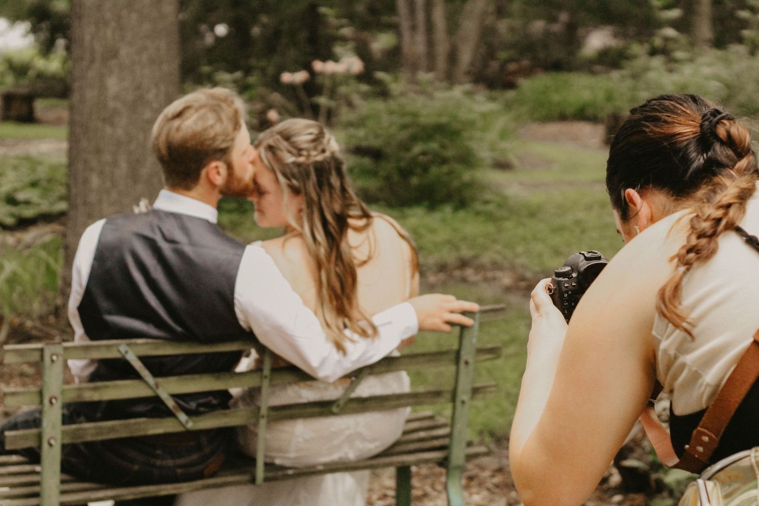 Wedding photographer capturing couple sitting on rustic bench in garden setting.