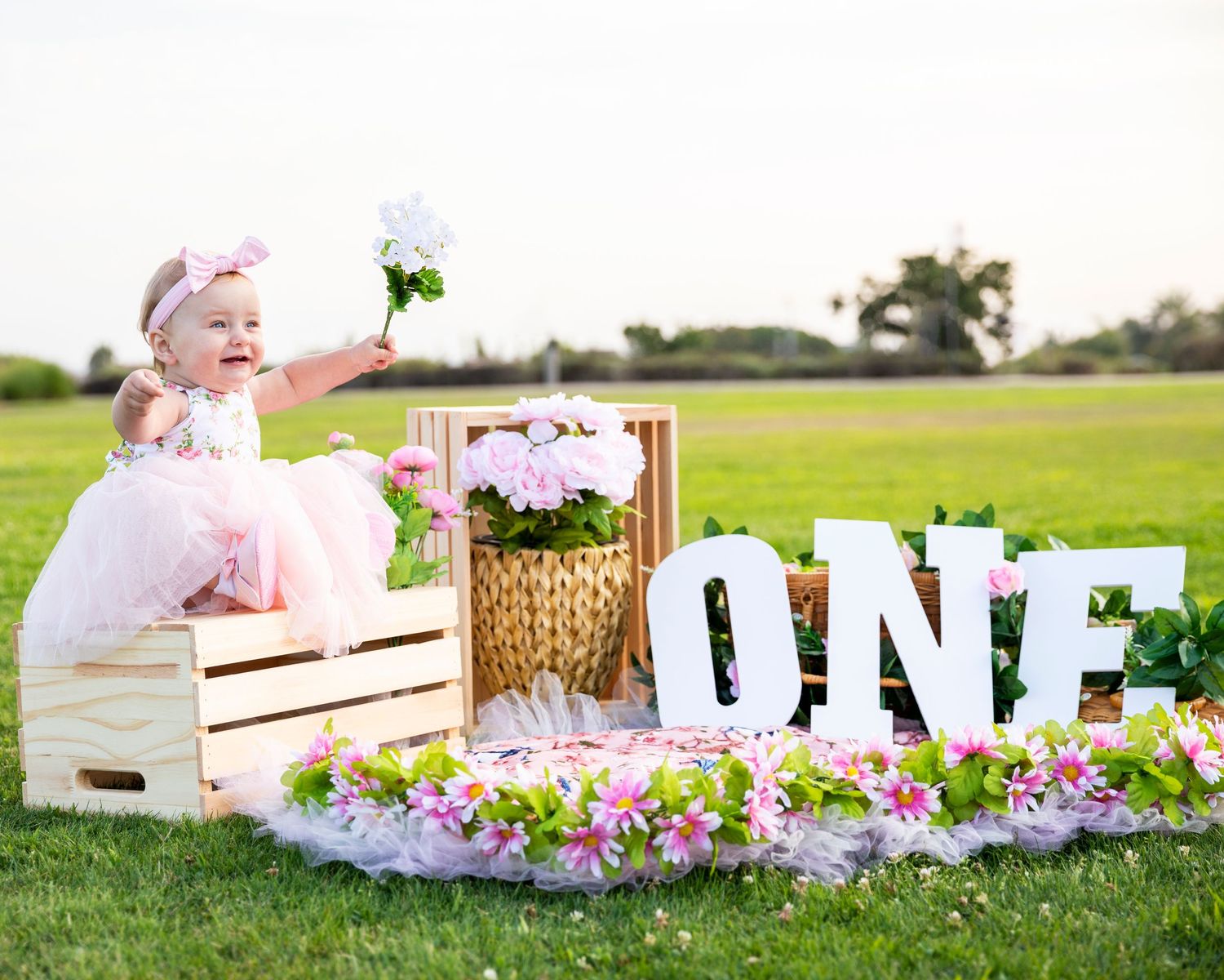 birthday photoshoot with Baby girl in pink dress sitting by ONE sign with flowers, holding a white bloom outdoors.