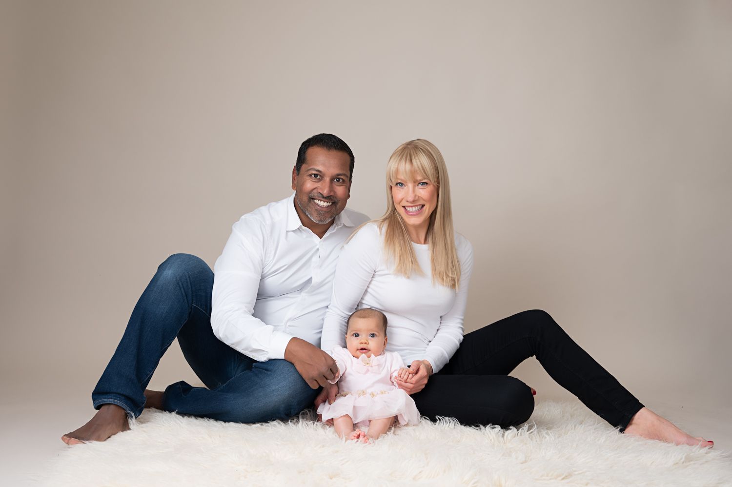 Family portrait on white fur rug featuring parents with baby in studio setting.
