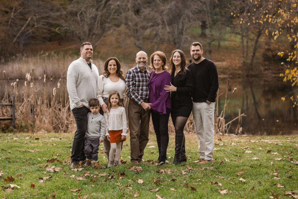 Extended family poses together on grass with autumn trees and a lake in the background.