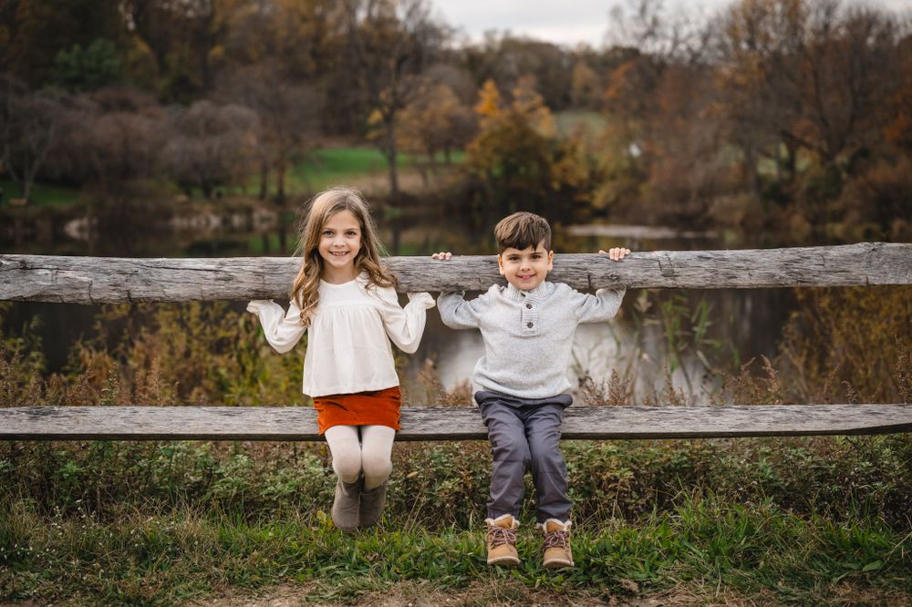 Children sit together on wooden fence during autumn outdoor photo session.