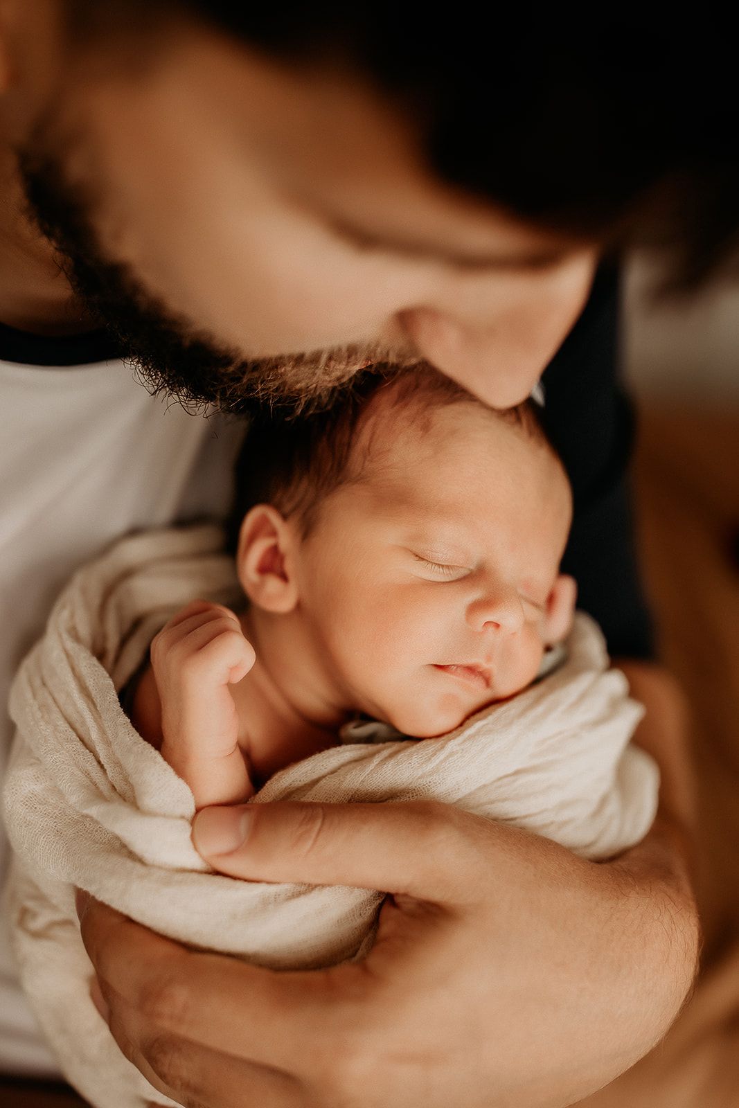A father leans down and kisses his swaddled newborn boy on the head.