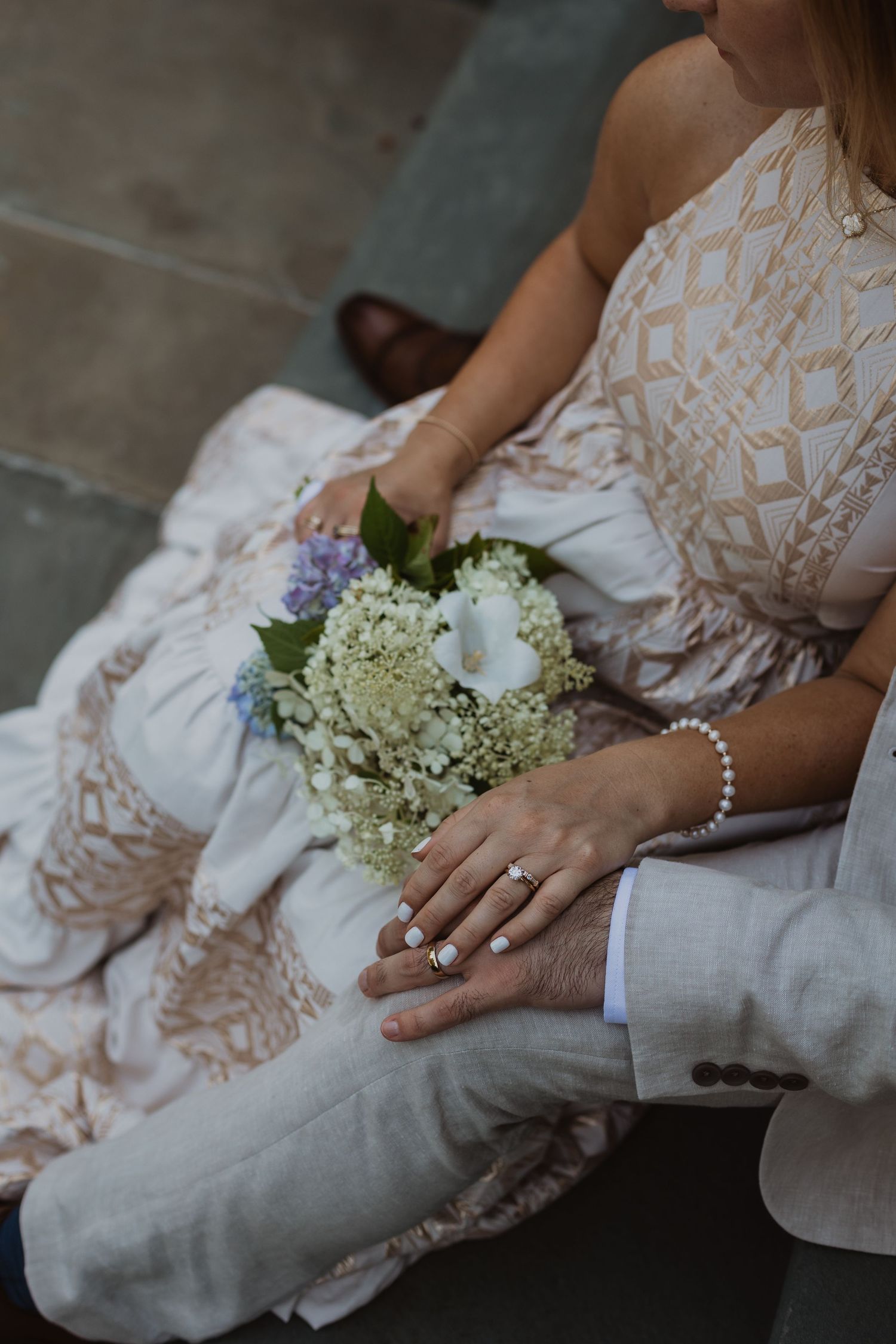 Close up of a wedding bouquet with white flowers and lace dress details as hands display rings and bracelet.