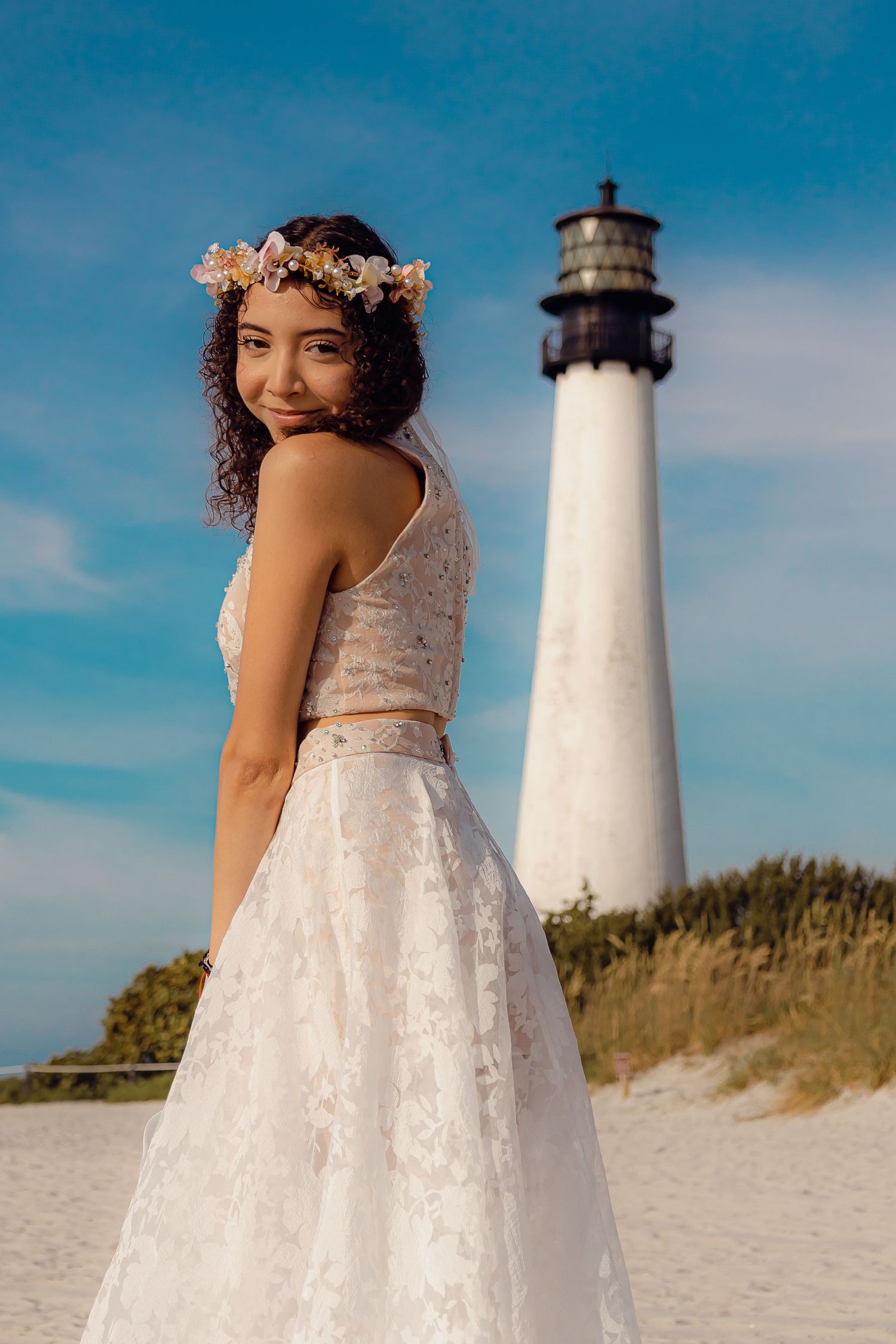 A white two-piece dress ensemble photographed in front of a coastal lighthouse on a sunny beach day.