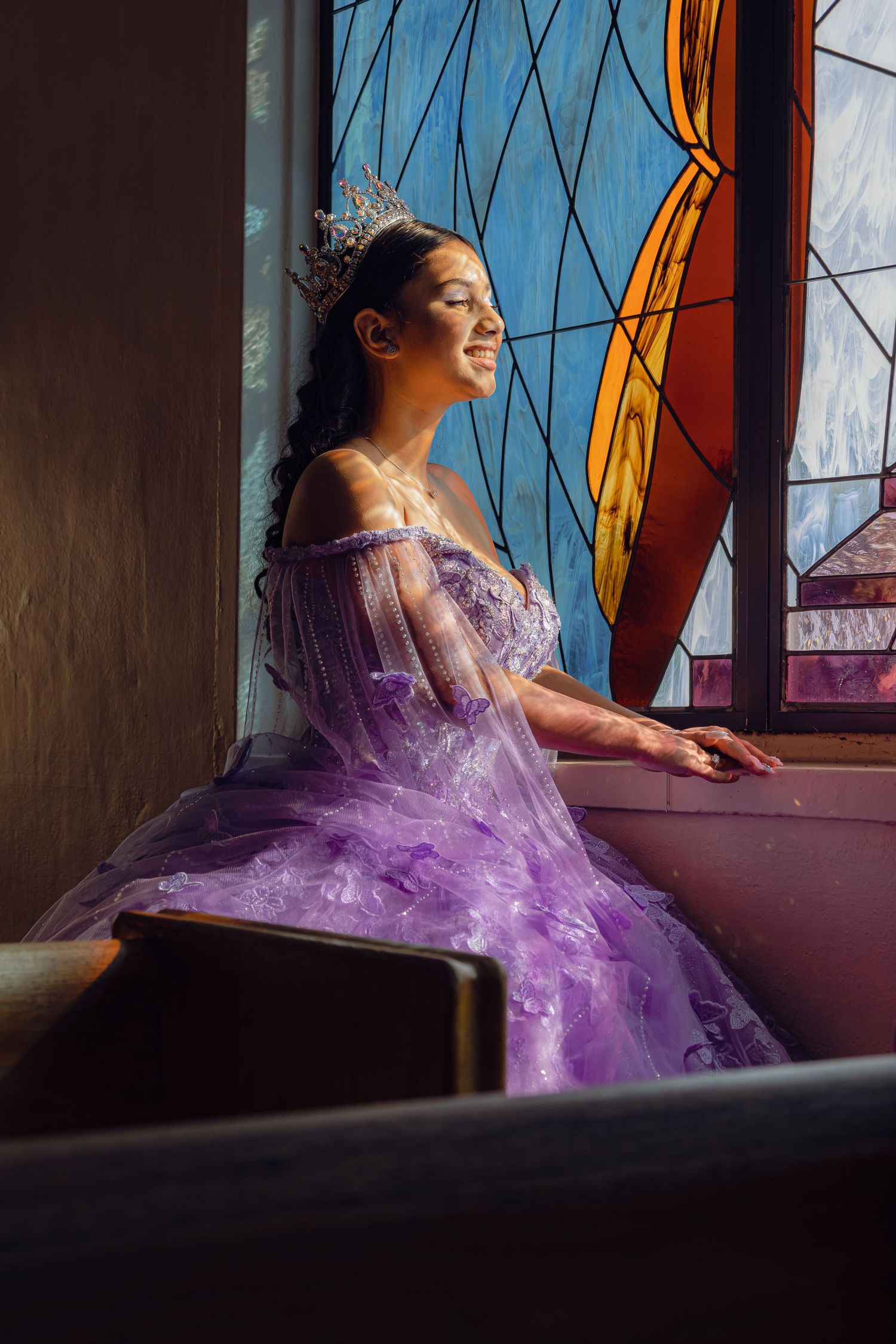 A dreamy portrait by stained glass windows featuring a purple princess gown and sparkling crown.