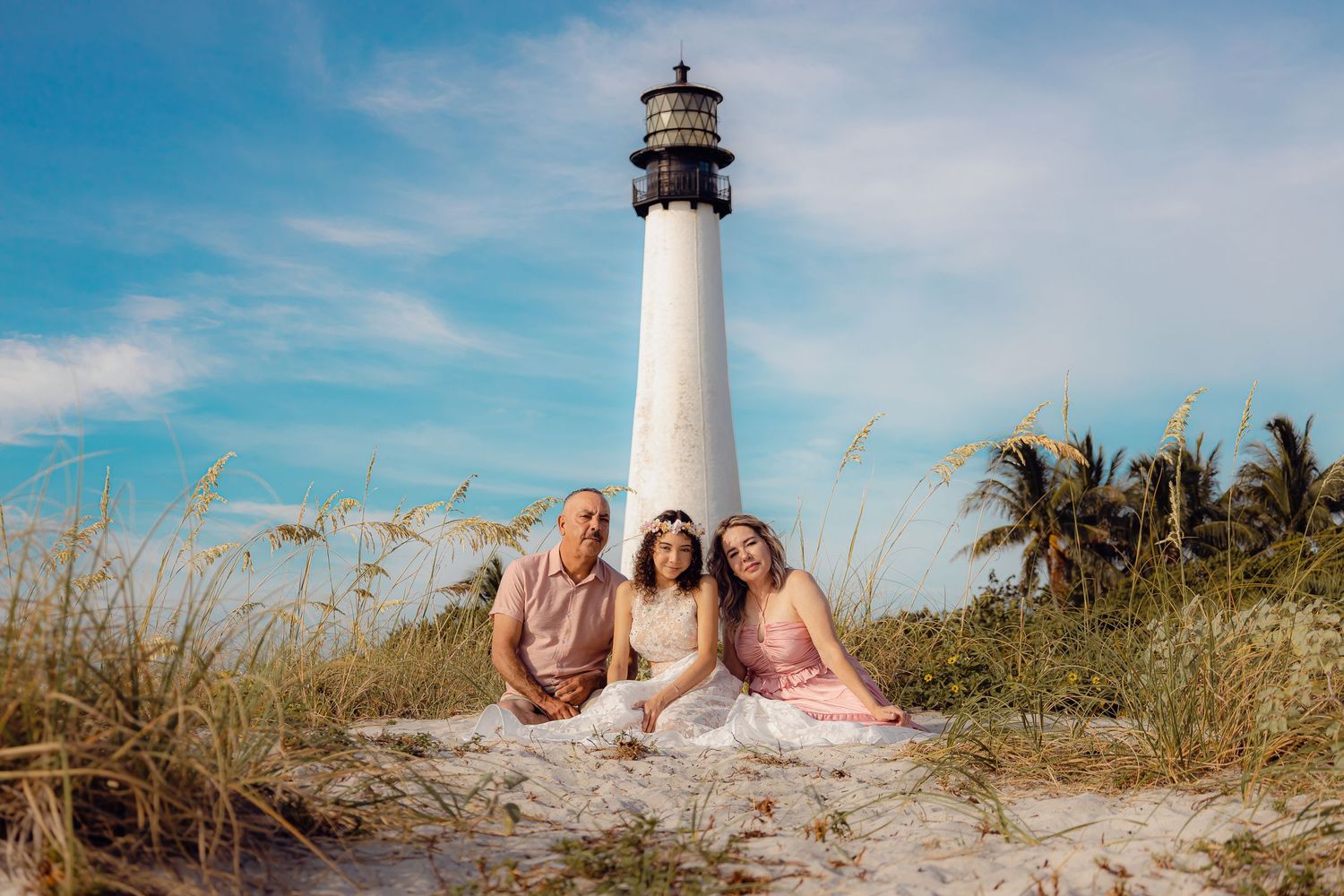 Three people in coordinated outfits pose on sand dunes with a lighthouse and tropical scenery.