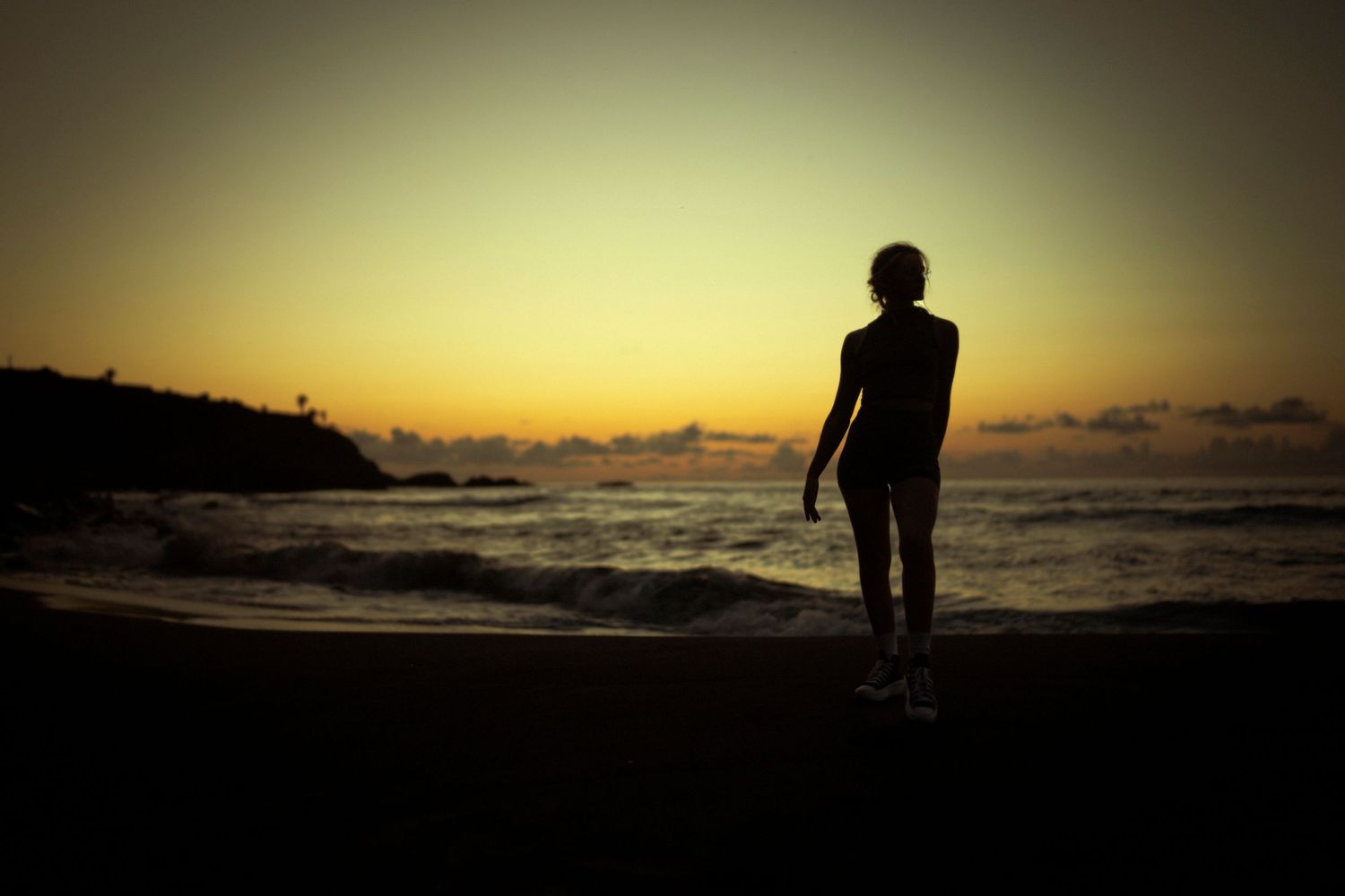 Silhouette of person standing on beach at sunset against orange sky.